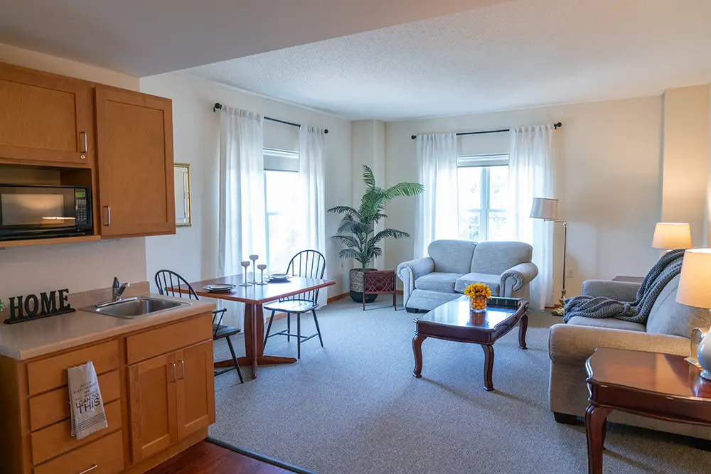 A cozy living and dining area at Sholom Senior Living features light-colored sofas, a wooden dining table with chairs, a small kitchen, large windows, white curtains, and indoor plants.