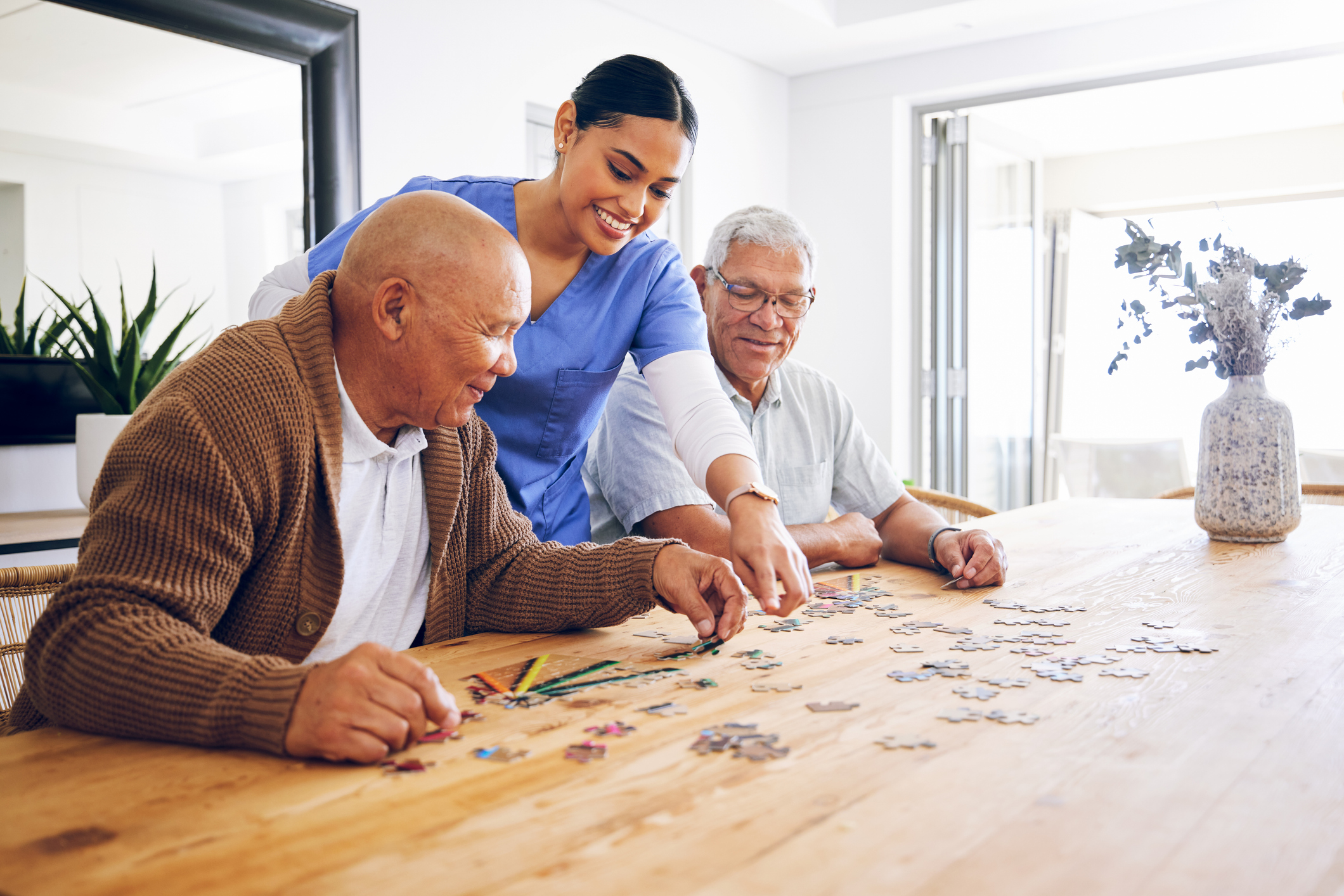 A caregiver at Sholom Senior Living helps two elderly men assemble a jigsaw puzzle at a wooden table in a bright, modern room filled with plants and a vase of flowers.