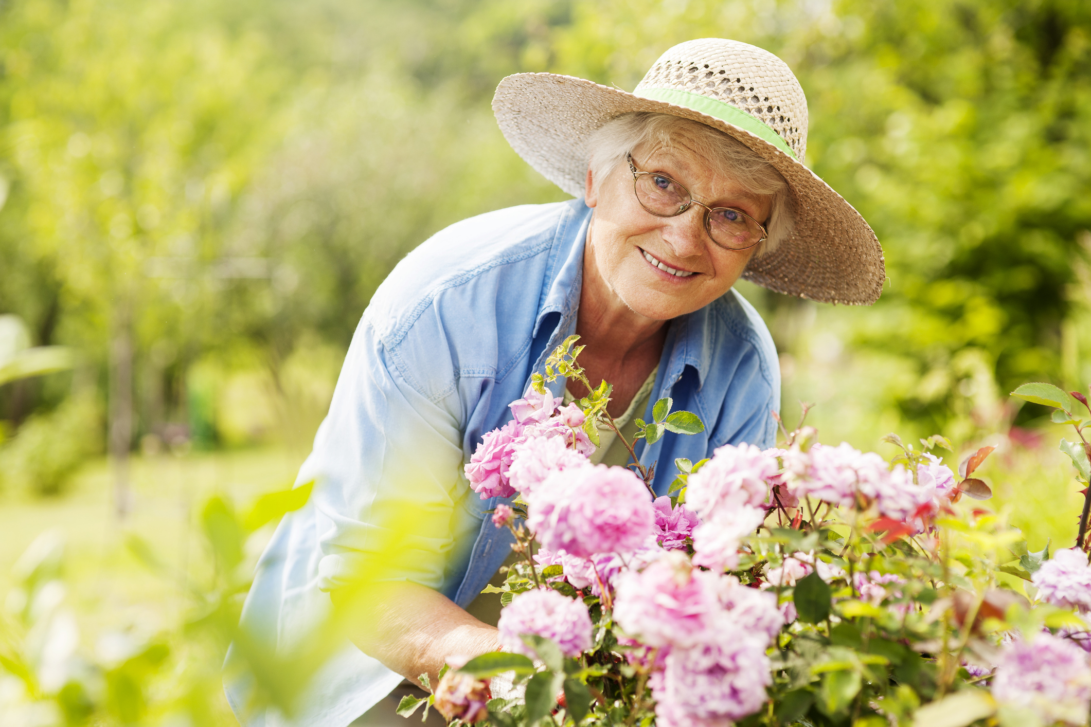 An older woman wearing a straw hat and glasses smiles while tending to pink flowers in a Sholom Senior Living garden.