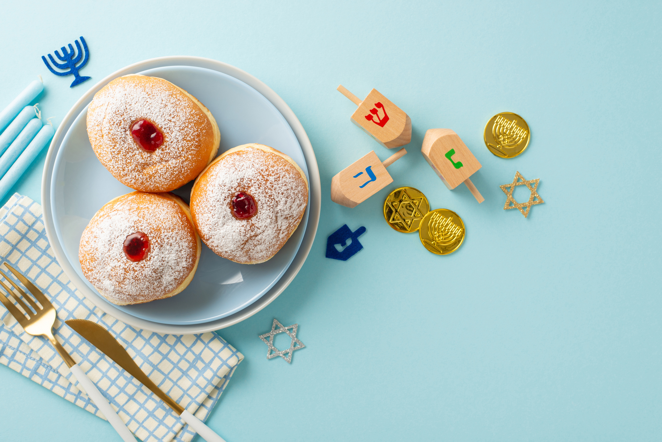 Three jelly-filled donuts on a plate with a napkin and cutlery, surrounded by Hanukkah candles, dreidels, chocolate coins, and Star of David decorations on a blue background at Sholom Senior Living.