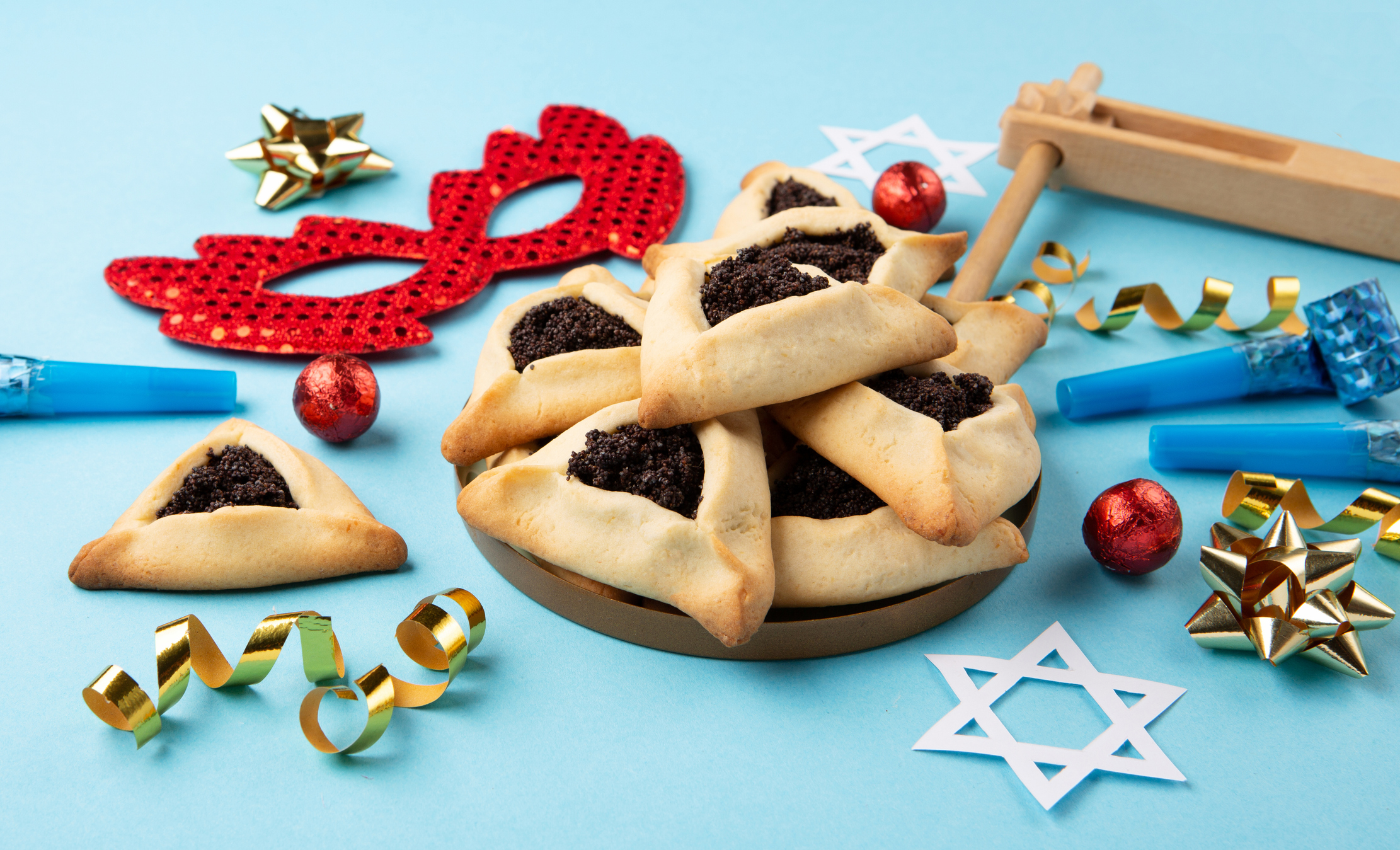 A plate of hamantaschen cookies surrounded by Purim decorations, including a mask, noisemaker, party blowers, star cutouts, and ribbons on a blue background at Sholom Senior Living.