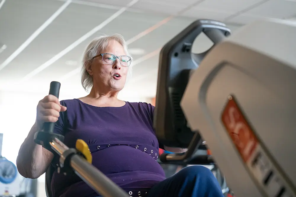 An older woman wearing glasses uses a recumbent exercise machine in the gym at Sholom Senior Living.