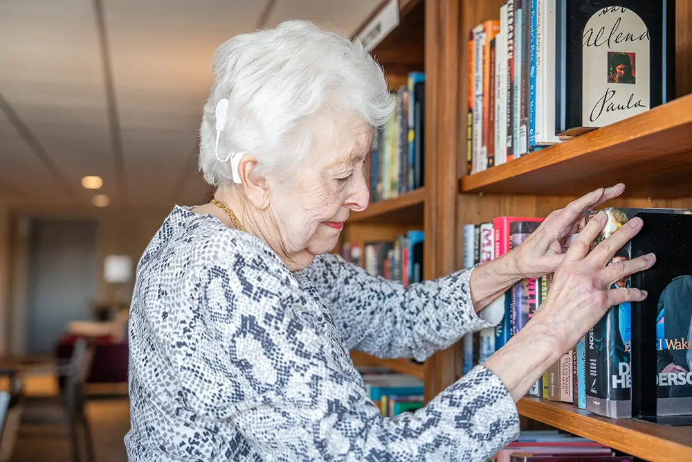 An elderly woman with white hair and a patterned blouse selects a book from a wooden bookshelf in a well-lit room at Sholom Senior Living.
