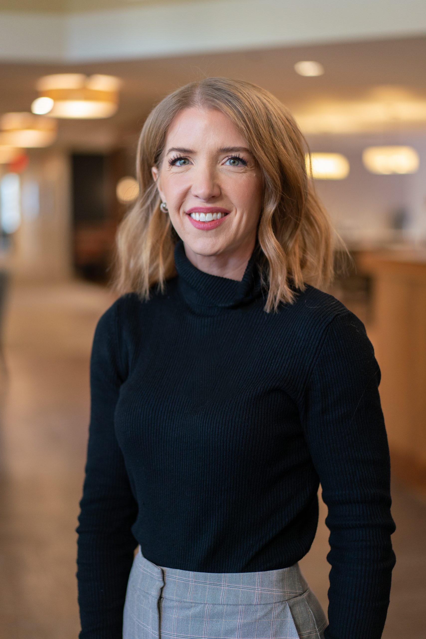 A woman with shoulder-length blonde hair, wearing a black turtleneck and plaid pants, stands indoors at Sholom Senior Living and smiles at the camera. A woman with shoulder-length blonde hair, wearing a black turtleneck and plaid pants, stands indoors at Sholom Senior Living and smiles at the camera.