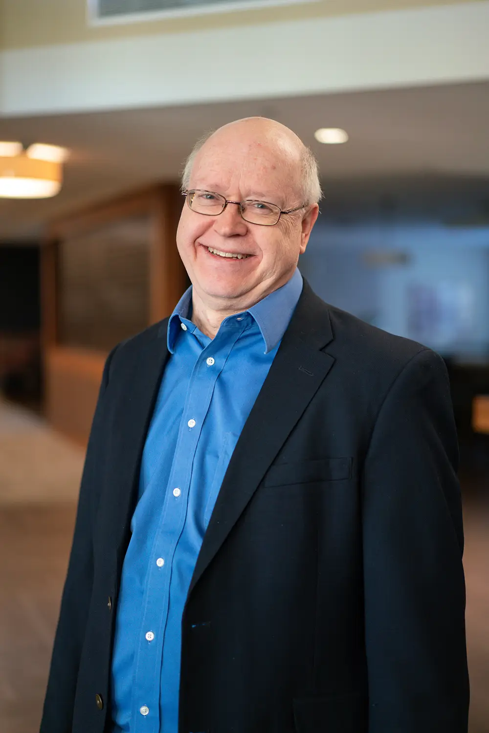 A man wearing glasses, a blue shirt, and a dark blazer stands indoors at Sholom Senior Living, smiling at the camera. The background is softly lit and out of focus. A man wearing glasses, a blue shirt, and a dark blazer stands indoors at Sholom Senior Living, smiling at the camera. The background is softly lit and out of focus.