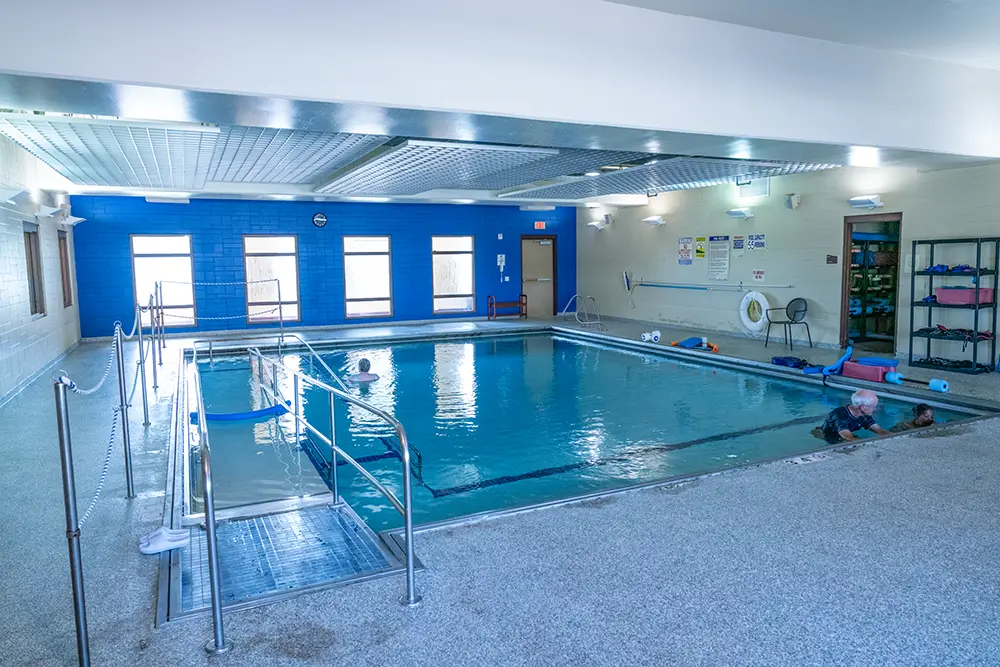 Indoor swimming pool with clear water, handrails, and pool equipment on shelves at Sholom Senior Living, with a few people enjoying the pool near the right side.