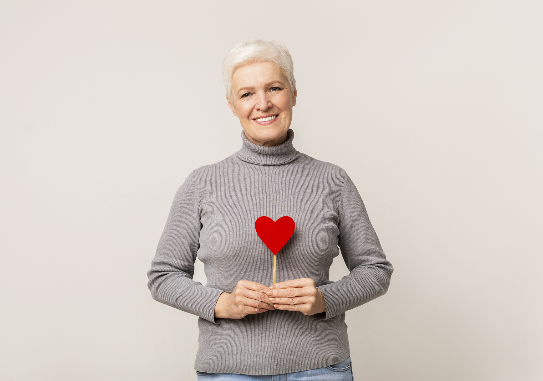Older woman with short white hair, wearing a gray turtleneck sweater, smiles while holding a red glittery heart on a stick against a plain light background—capturing the warmth of Sholom Senior Living.