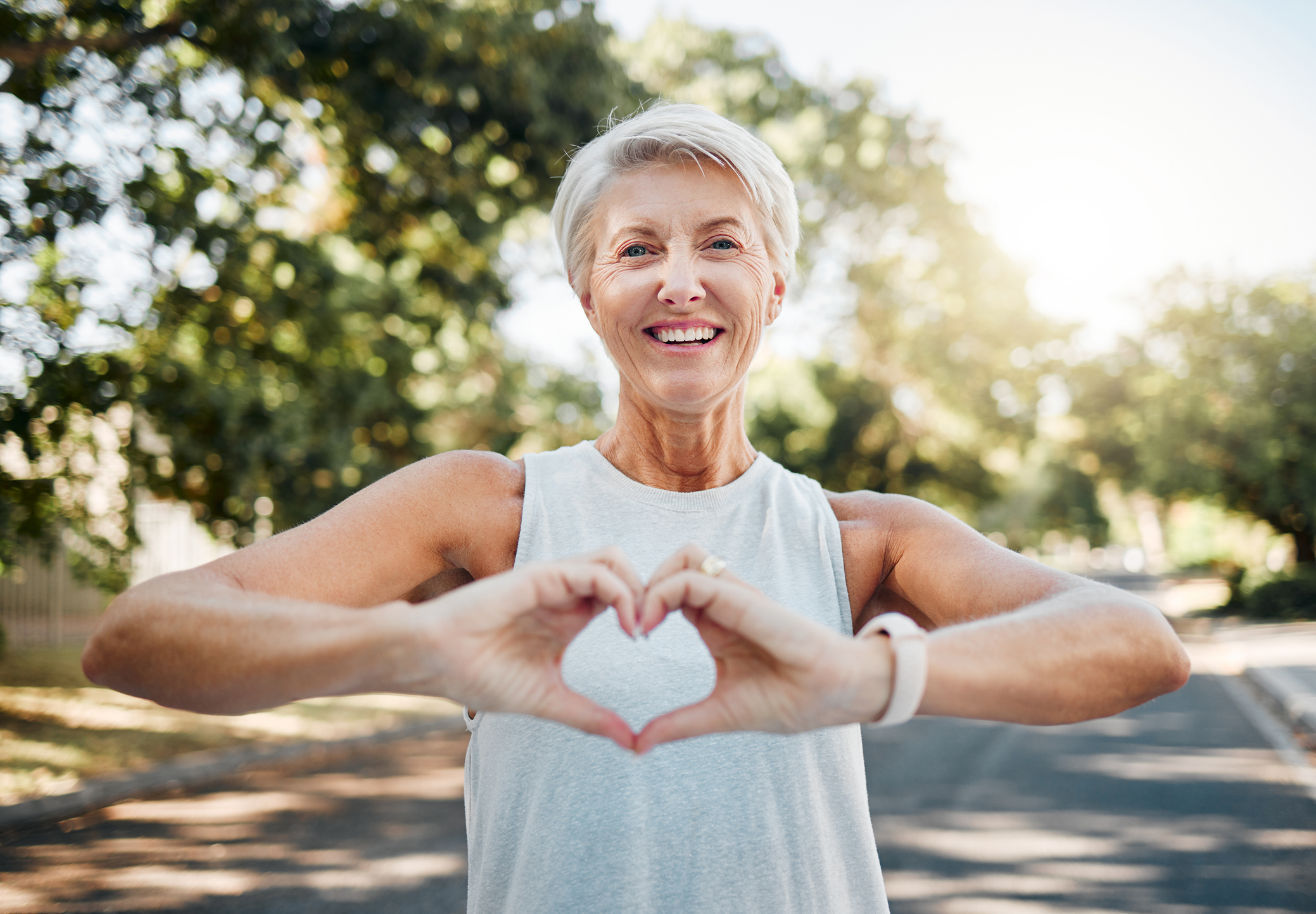 Smiling older woman outside at Sholom Senior Living, making a heart shape with her hands while wearing a sleeveless top, with trees and sunlight in the background. Smiling older woman outside at Sholom Senior Living, making a heart shape with her hands while wearing a sleeveless top, with trees and sunlight in the background.