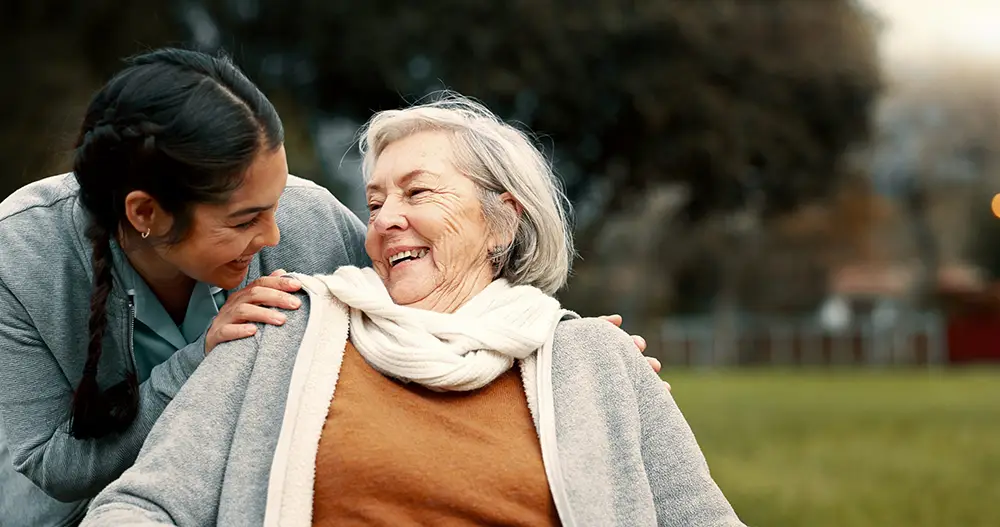 A younger woman smiles and puts her hand on the shoulder of an older woman in a wheelchair. Both are outdoors on a grassy area, enjoying each other’s company at Sholom Senior Living.
