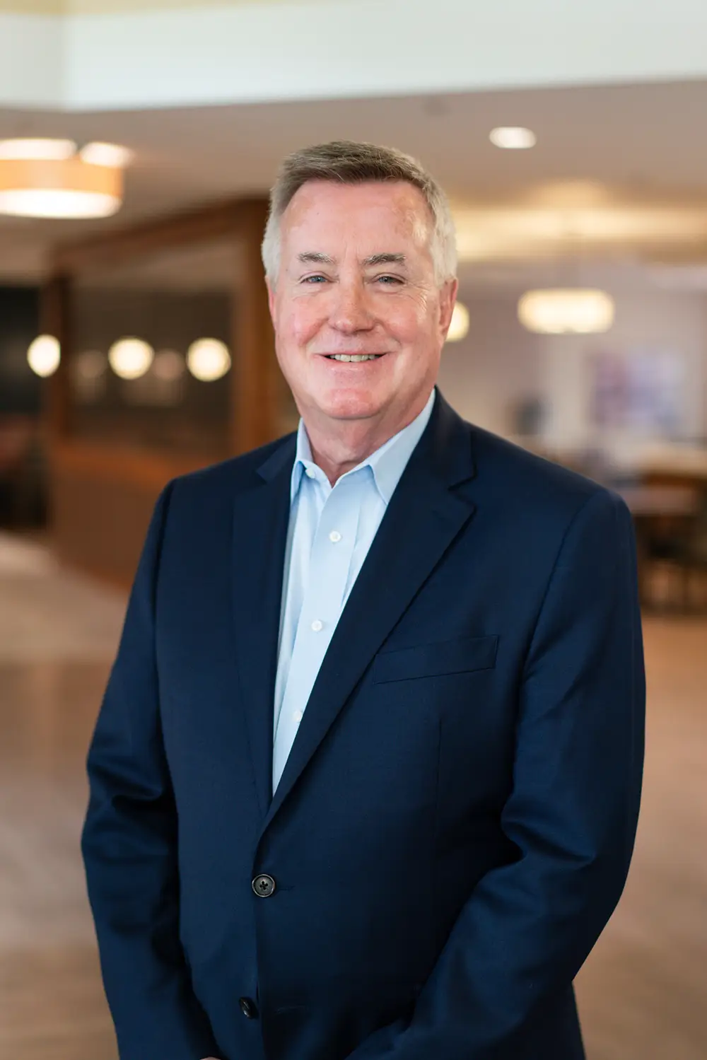 A middle-aged man in a navy suit and light blue shirt stands indoors, smiling at the camera. The background shows a modern, blurred Sholom Senior Living office setting. A middle-aged man in a navy suit and light blue shirt stands indoors, smiling at the camera. The background shows a modern, blurred Sholom Senior Living office setting.