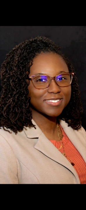 A woman with curly hair wearing glasses, a beige blazer, and an orange top, smiles in front of a black background at Sholom Senior Living. A woman with curly hair wearing glasses, a beige blazer, and an orange top, smiles in front of a black background at Sholom Senior Living.