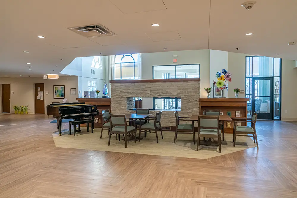 Spacious communal area at Sholom Senior Living with a piano, grouped chairs and tables, a double-sided fireplace, and large windows letting in natural light.