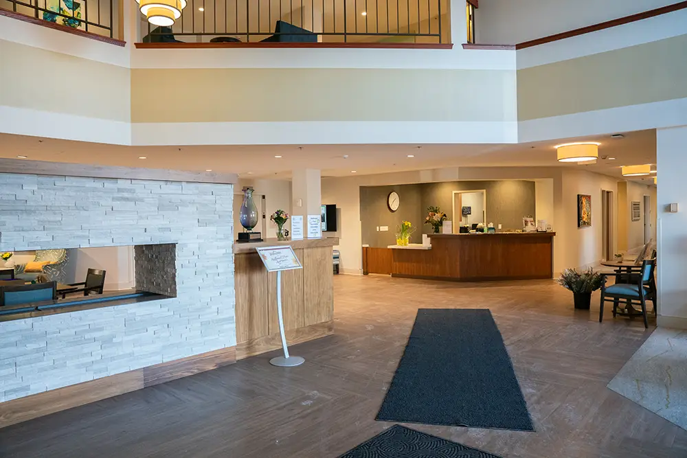 A hotel lobby with a reception desk, modern furnishings, a stone accent wall, chairs, and signage on a stand welcomes guests to Sholom Senior Living.