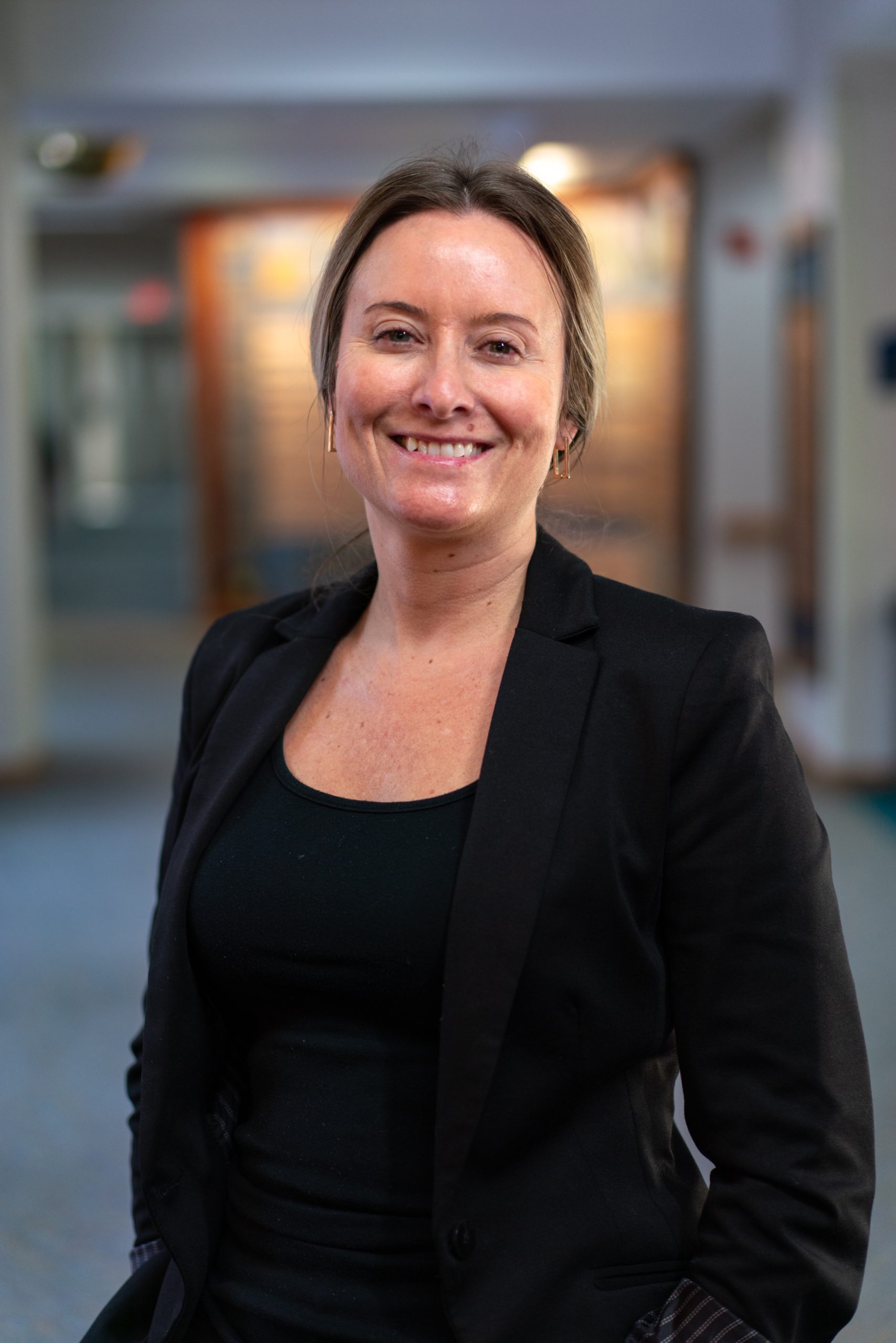 A woman with light hair tied back, wearing a black blazer and top, stands indoors at Sholom Senior Living and smiles at the camera. A woman with light hair tied back, wearing a black blazer and top, stands indoors at Sholom Senior Living and smiles at the camera.