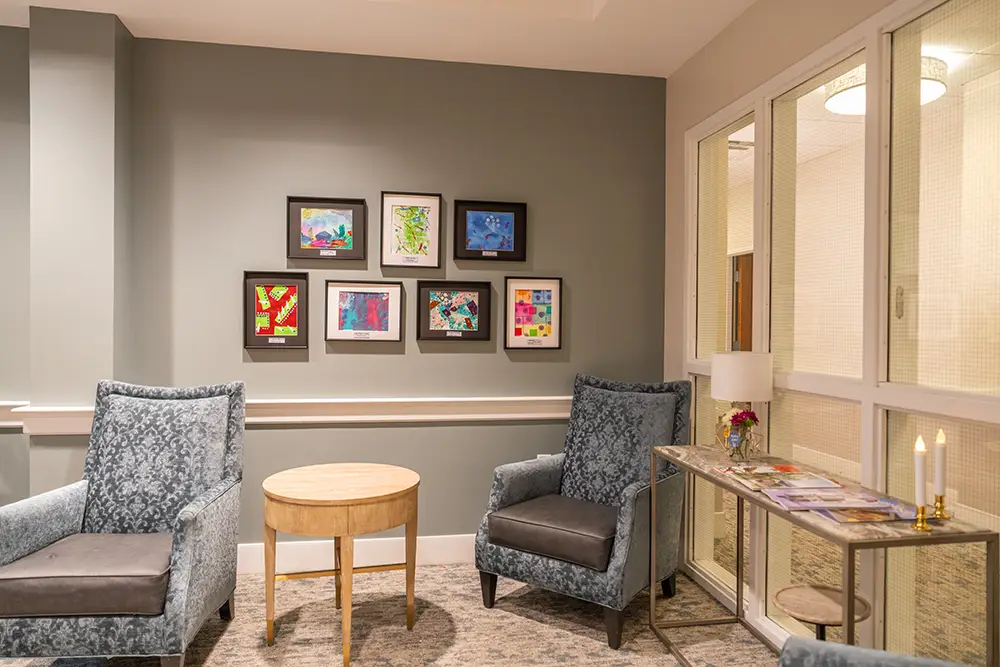 Two patterned armchairs and a round wooden table sit in a cozy corner at Sholom Senior Living; children's artwork brightens the gray wall, while a console table with magazines and a lamp stands by the glass partition.
