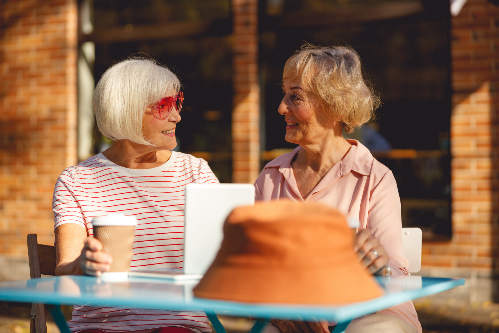 Two older women sit at an outdoor table at Sholom Senior Living, chatting and smiling over coffee and a tablet. A brown hat rests on the table, with a brick building in the background.