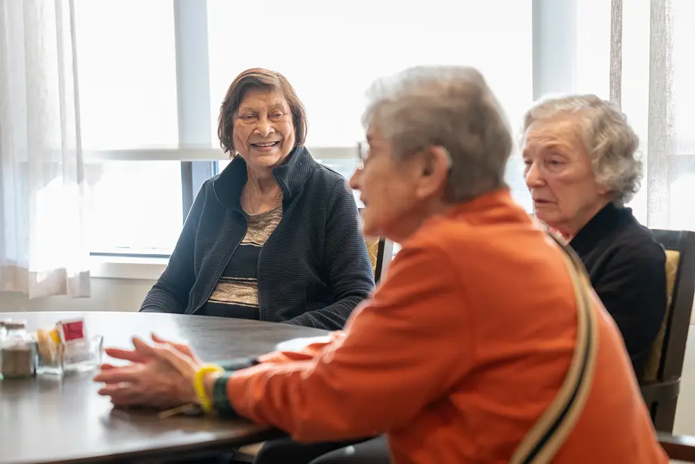 Three elderly women sit around a table in a bright room at Sholom Senior Living, engaged in conversation. Three elderly women sit around a table in a bright room at Sholom Senior Living, engaged in conversation.