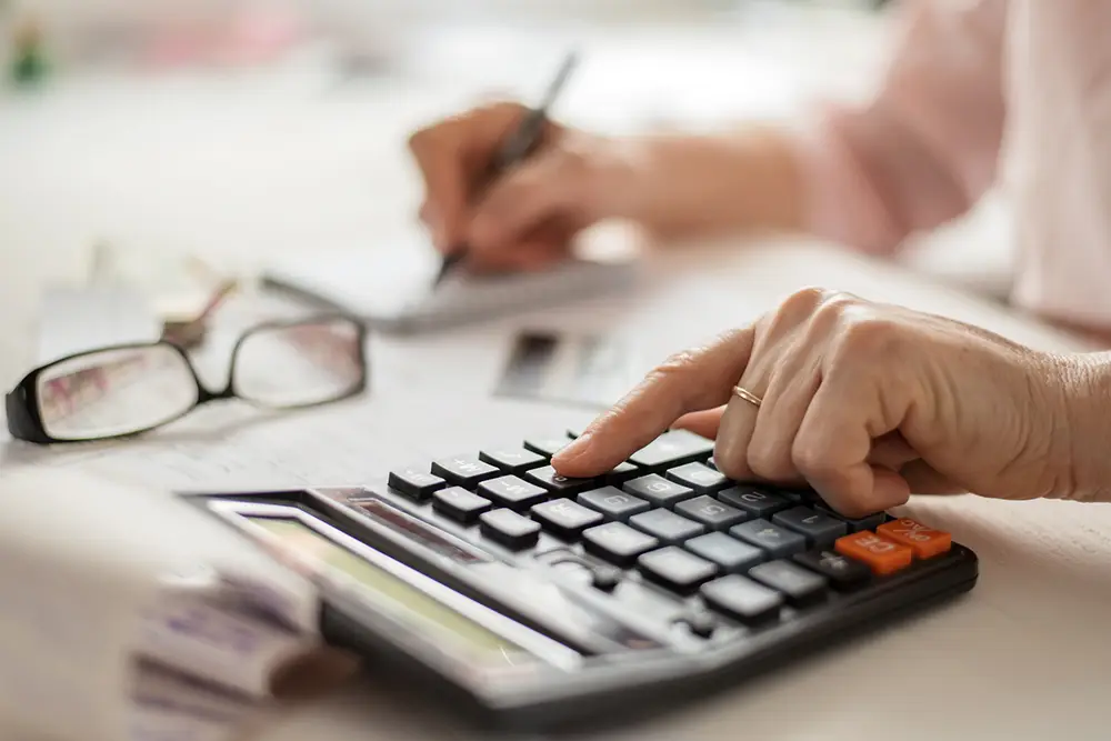 A person at a desk uses a calculator while writing on paper, surrounded by eyeglasses and documents—capturing a moment of organization at Sholom Senior Living. A person at a desk uses a calculator while writing on paper, surrounded by eyeglasses and documents—capturing a moment of organization at Sholom Senior Living.