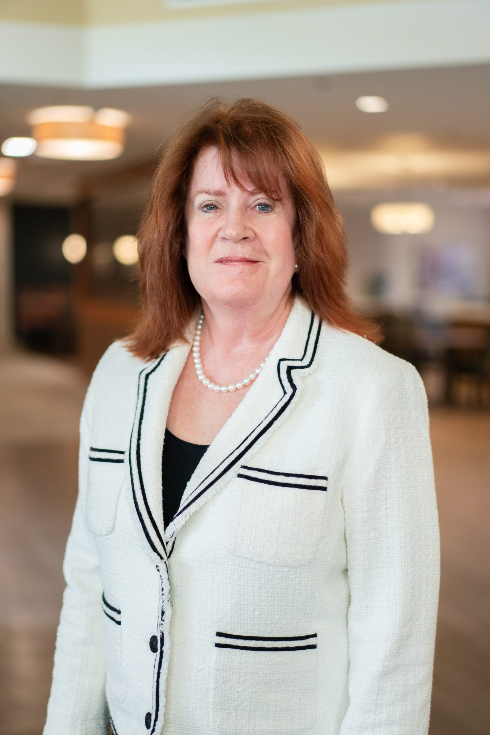 Woman with shoulder-length auburn hair wearing a white blazer with black trim and a pearl necklace, standing indoors in the well-lit lobby of Sholom Senior Living. Woman with shoulder-length auburn hair wearing a white blazer with black trim and a pearl necklace, standing indoors in the well-lit lobby of Sholom Senior Living.