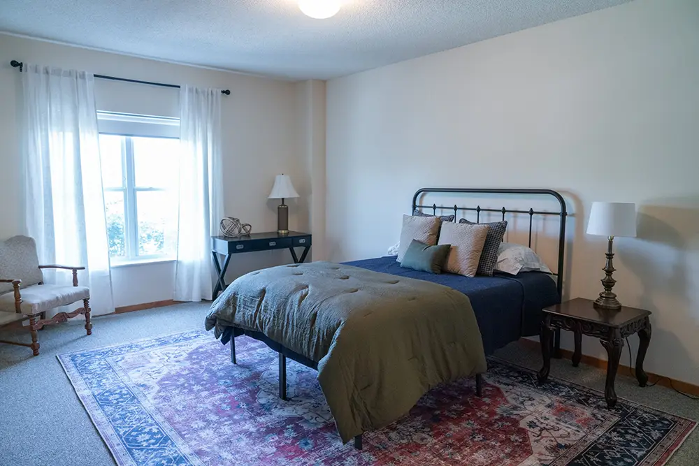A cozy bedroom at Sholom Senior Living features a double bed with pillows and a green comforter, two side tables with lamps, a chair, a desk by the window, and a patterned rug on the floor. A cozy bedroom at Sholom Senior Living features a double bed with pillows and a green comforter, two side tables with lamps, a chair, a desk by the window, and a patterned rug on the floor.