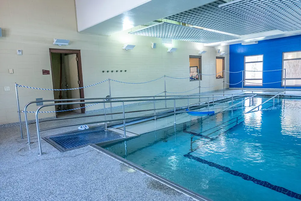 An indoor swimming pool at Sholom Senior Living features a metal accessibility ramp, blue wall, large windows, and a rope barrier separating the ramp from the pool area. An indoor swimming pool at Sholom Senior Living features a metal accessibility ramp, blue wall, large windows, and a rope barrier separating the ramp from the pool area.