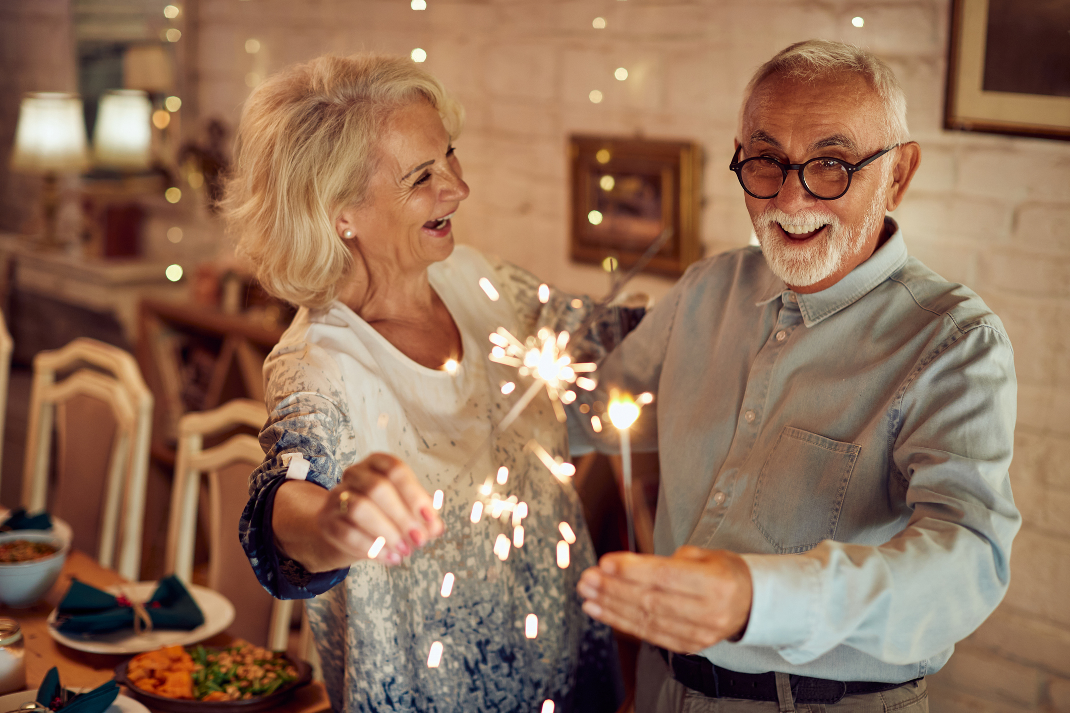Older couple indoors holding sparklers and smiling, surrounded by a festive setting with a dining table in the background at Sholom Senior Living.