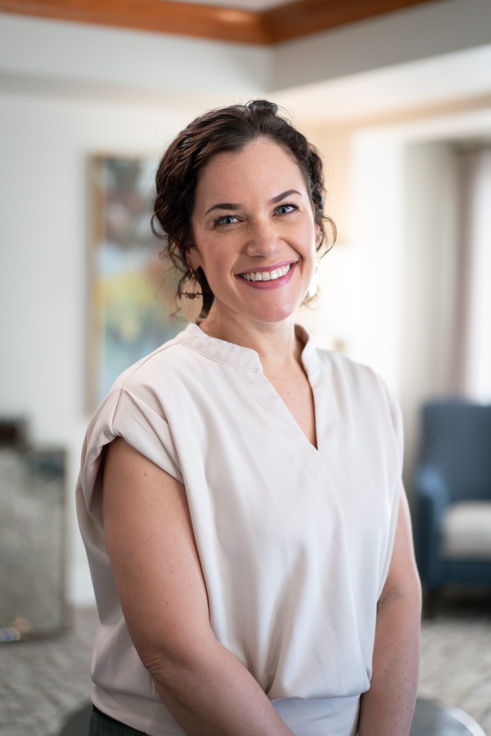 A woman with curly hair, wearing a light-colored blouse, stands indoors at Sholom Senior Living, smiling at the camera. The background features blurred home furnishings. A woman with curly hair, wearing a light-colored blouse, stands indoors at Sholom Senior Living, smiling at the camera. The background features blurred home furnishings.