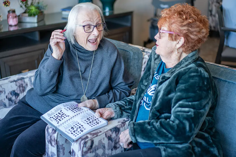 Two elderly women sit on a couch at Sholom Senior Living, one holding a pen and a crossword puzzle book. They engage in cheerful conversation, sharing laughter and enjoying each other's company.
