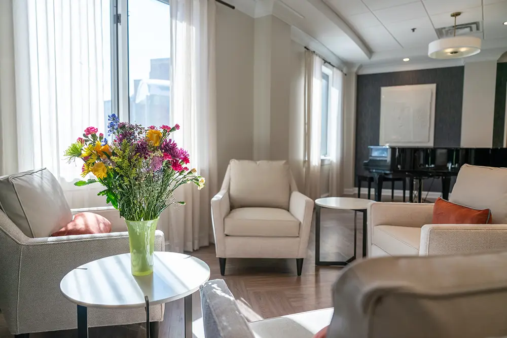 A bright living room at Sholom Senior Living features beige armchairs, a round side table with a vase of colorful flowers, large windows with white curtains, and a black grand piano in the background.