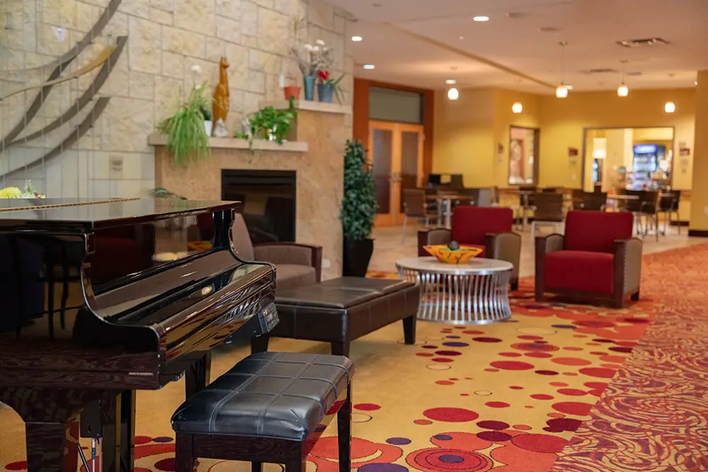 The Sholom Senior Living hotel lobby features a grand piano with a black bench, red and brown chairs, a cozy fireplace, and a dining area in the background. The carpet showcases an elegant red and yellow circular pattern.