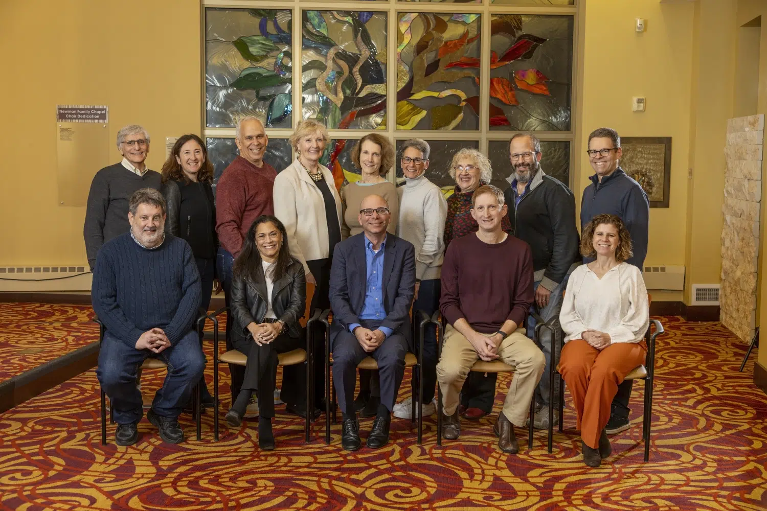 A group of fifteen adults at Sholom Senior Living pose indoors in front of a colorful stained glass window, with some seated in chairs and others standing behind them. A group of fifteen adults at Sholom Senior Living pose indoors in front of a colorful stained glass window, with some seated in chairs and others standing behind them.