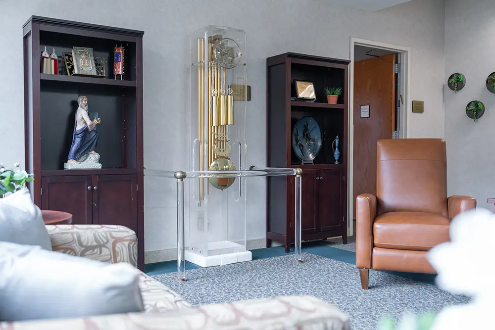 A welcoming waiting room at Sholom Senior Living features a brown armchair, patterned sofa, two wooden cabinets, a large glass-encased clock, and decorative items on display.