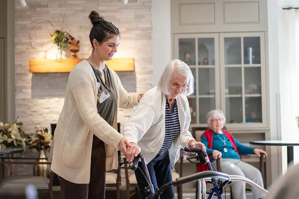 A caregiver assists an elderly woman using a walker at Sholom Senior Living, while another elderly woman sits in the background of the common room.