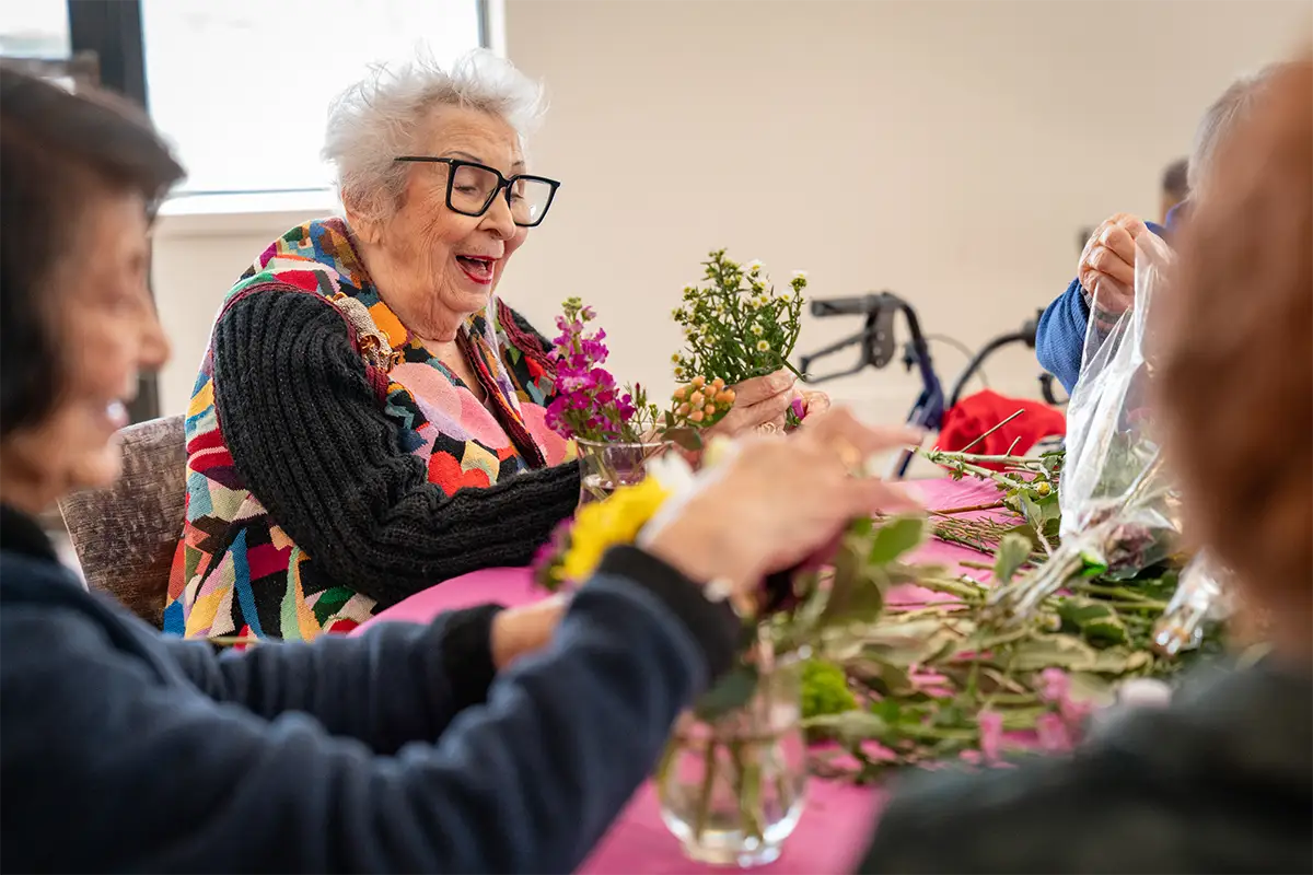 Several elderly women at Sholom Senior Living sit around a table arranging flowers, with one woman smiling and laughing while holding a bouquet. Several elderly women at Sholom Senior Living sit around a table arranging flowers, with one woman smiling and laughing while holding a bouquet.