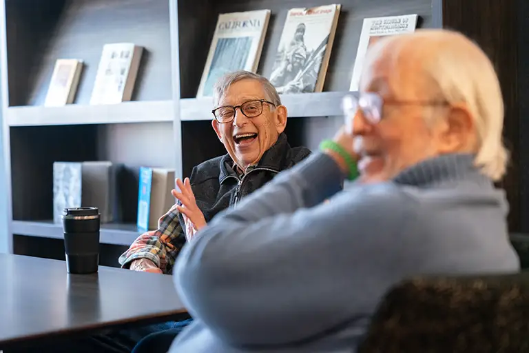 Two older men sit at a table in front of bookshelves, smiling and talking at Sholom Senior Living. One man is in focus, while the other is blurred in the foreground. Two older men sit at a table in front of bookshelves, smiling and talking at Sholom Senior Living. One man is in focus, while the other is blurred in the foreground.