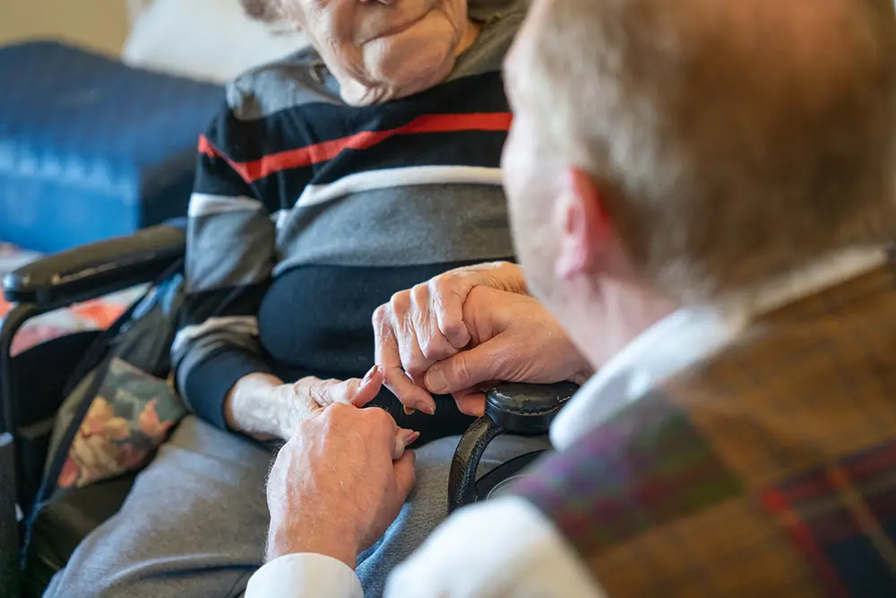 An elderly person in a wheelchair holds hands with another, offering comfort and support at Sholom Senior Living.