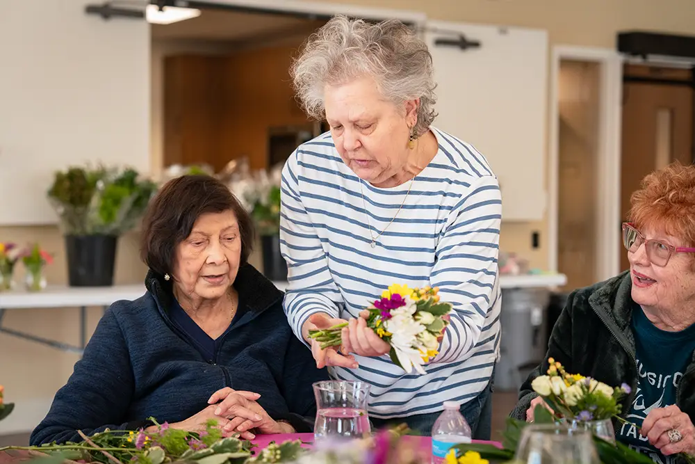 Three elderly women sit at a table making floral arrangements at Sholom Senior Living; one arranges flowers while the others watch and participate. Three elderly women sit at a table making floral arrangements at Sholom Senior Living; one arranges flowers while the others watch and participate.
