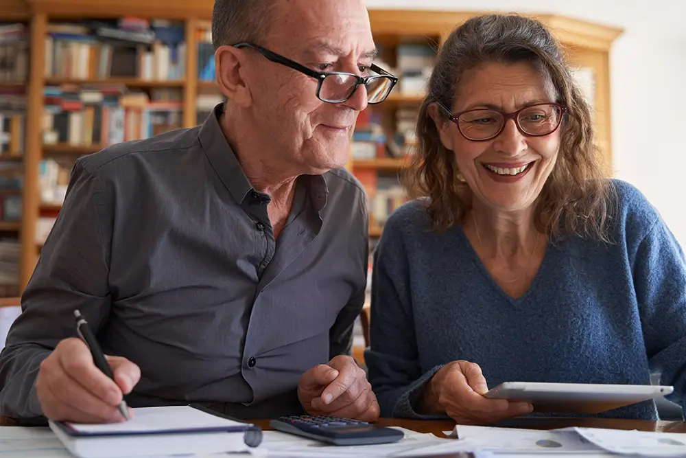 Two older adults sit at a table with papers, a calculator, a notebook, and a tablet, reviewing finances or documents together in a cozy Sholom Senior Living home office or library.