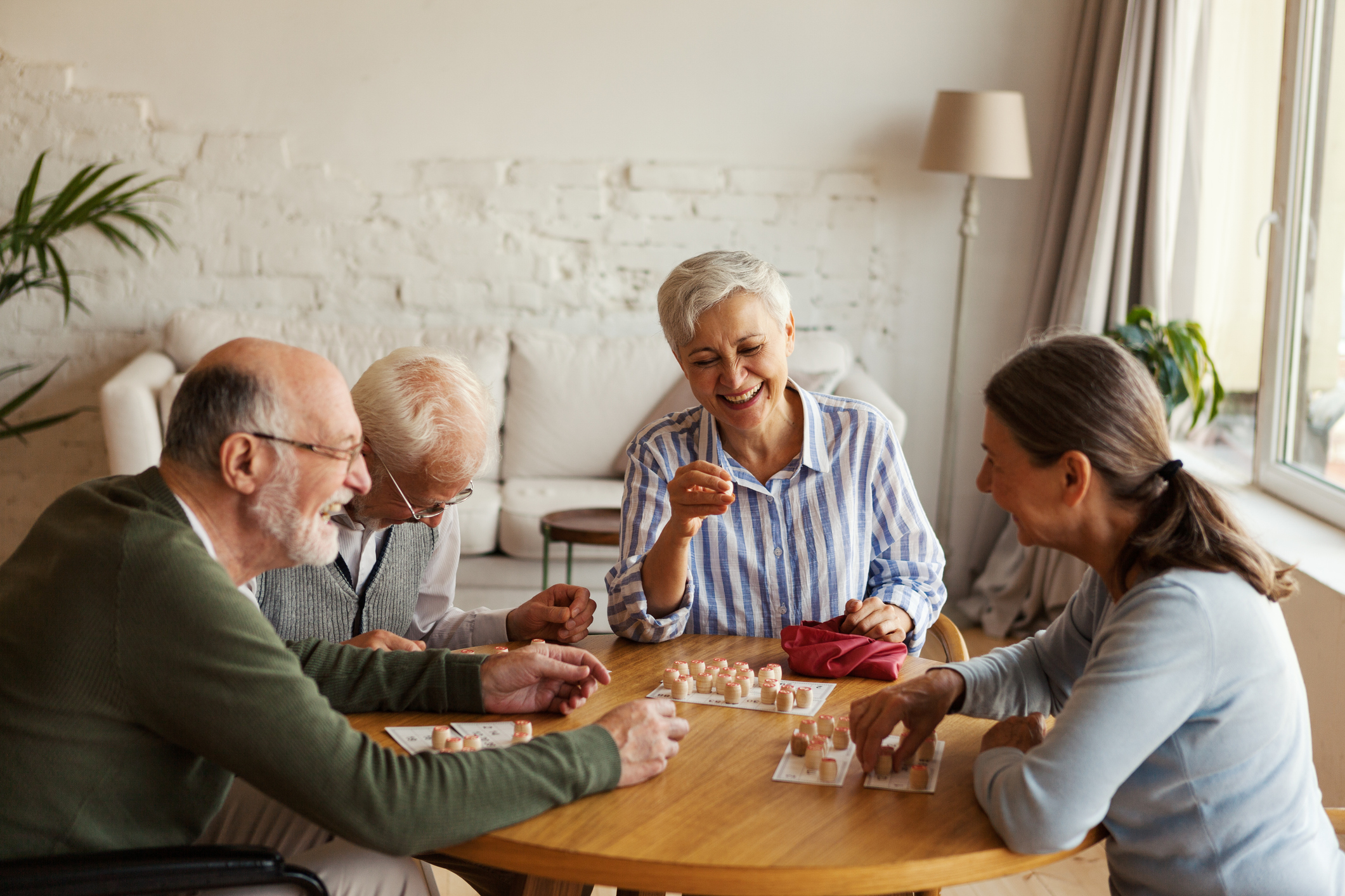 Four older adults sit around a table at Sholom Senior Living, playing a board game while smiling and laughing in a well-lit living room.
