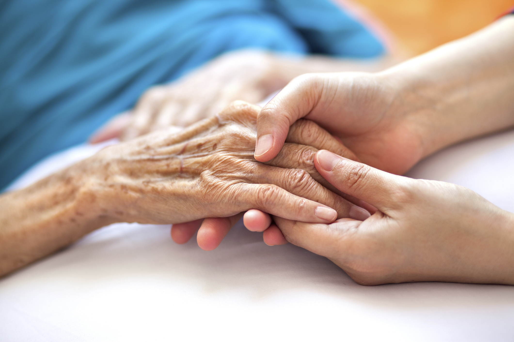 A younger person gently holds the hand of an elderly person lying in bed at Sholom Senior Living, showing care and support.