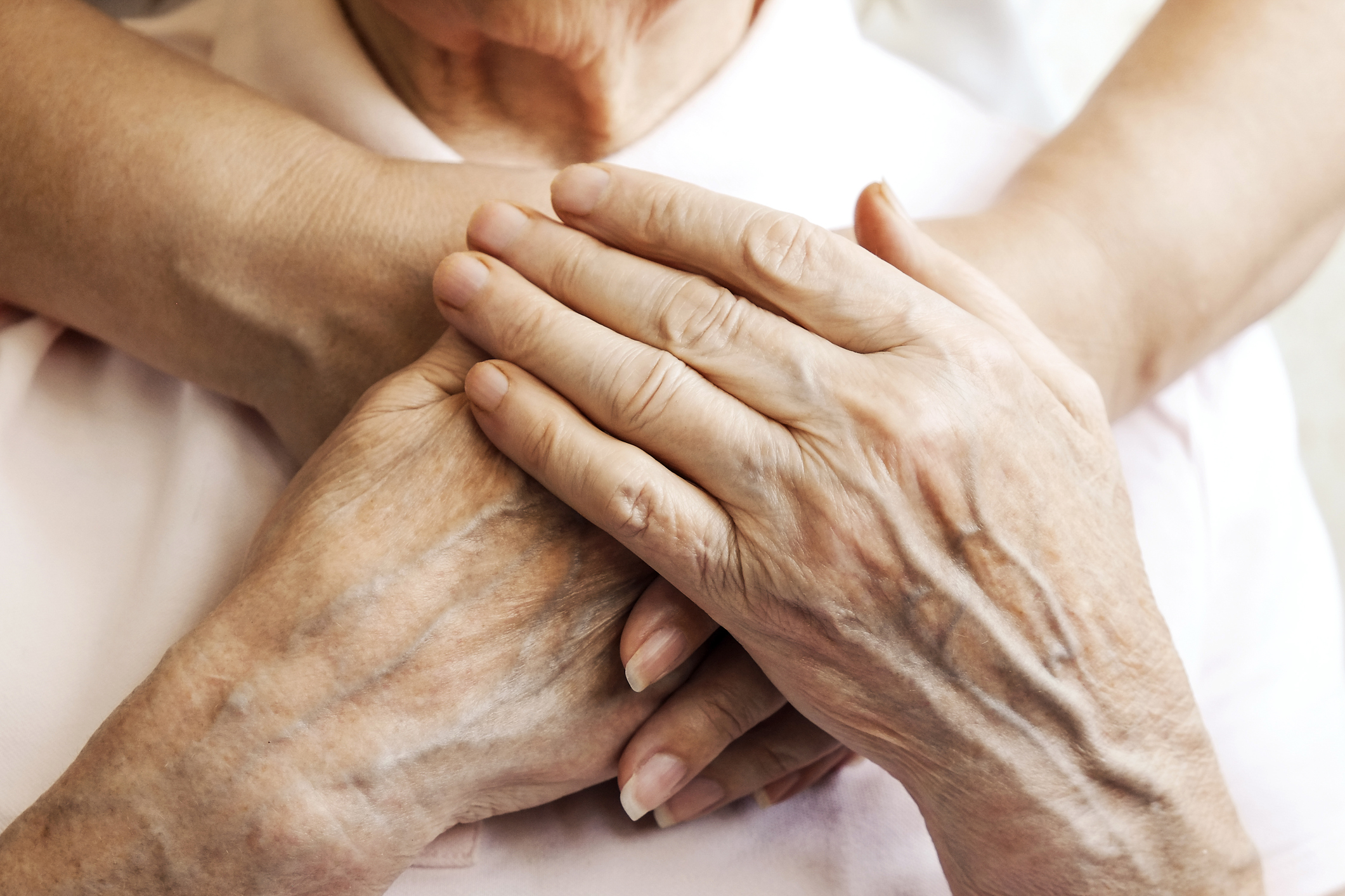 Close-up of an elderly person’s hands resting on their chest, with another person's arms gently embracing them from behind at Sholom Senior Living.