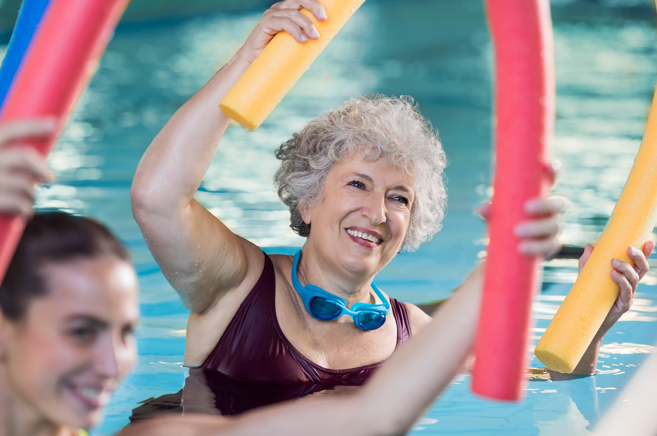 An older woman with short gray hair in a swimming pool at Sholom Senior Living, holding a yellow pool noodle and goggles around her neck, participates in a group aquatic exercise.