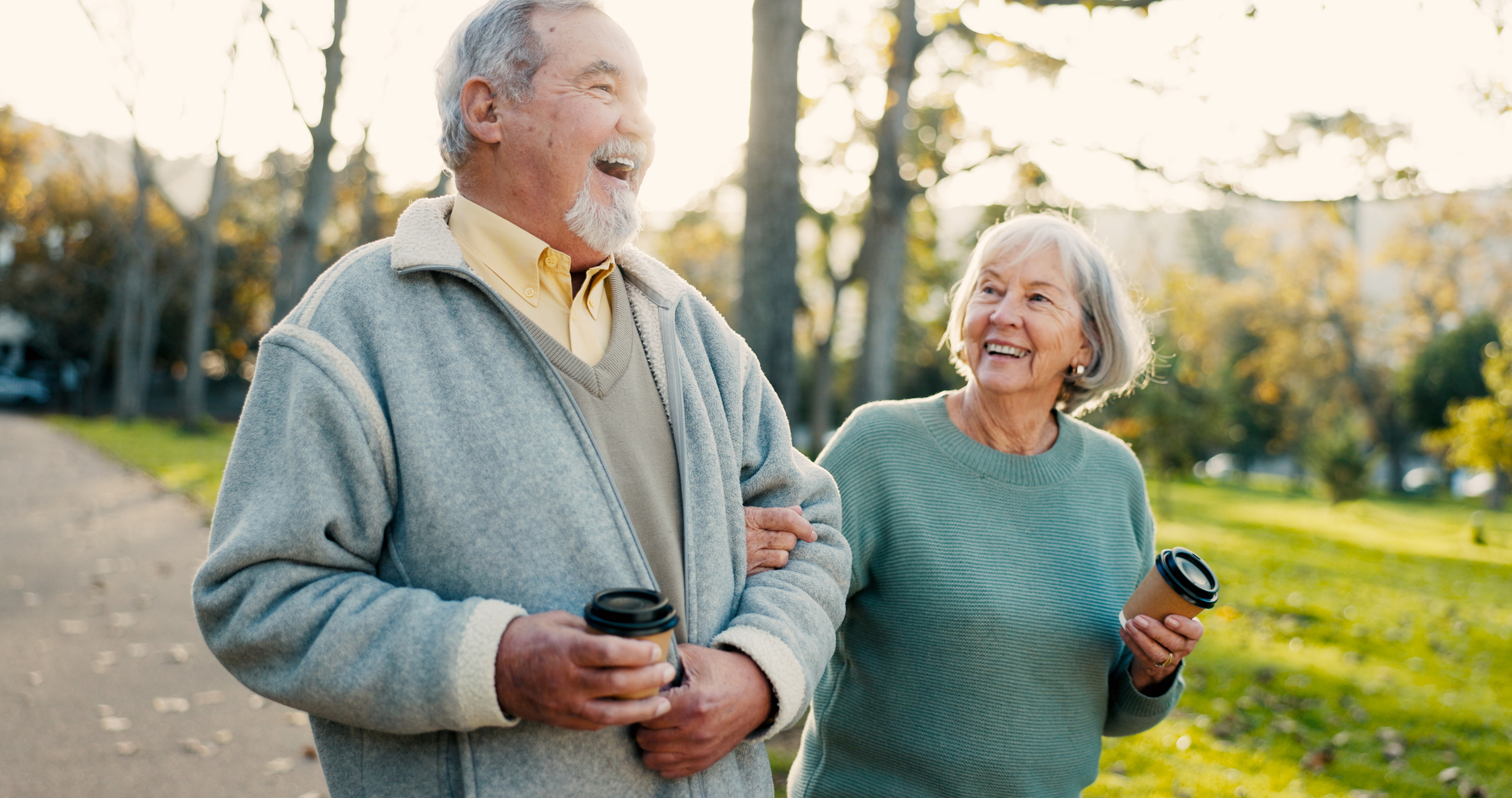 An older man and woman walk arm in arm through a park, smiling and holding coffee cups on a sunny day, enjoying the community spirit of Sholom Senior Living.