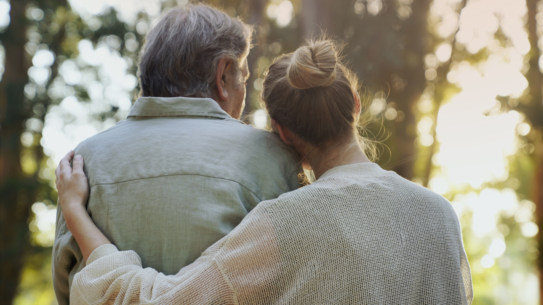 Two people are seen from behind; one has an arm around the other, enjoying the outdoors at Sholom Senior Living with sunlight and trees in the background.