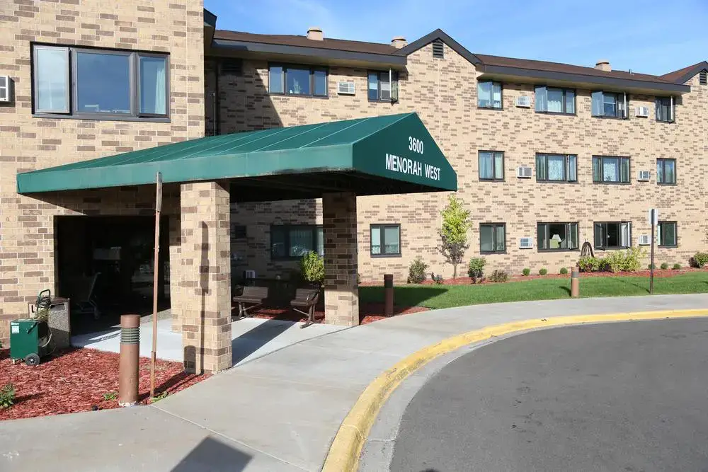 The entrance to a brick apartment building, part of Sholom Senior Living, features a green canopy reading "3300 MENORAH WEST"; landscaped grounds and a curved driveway are visible.