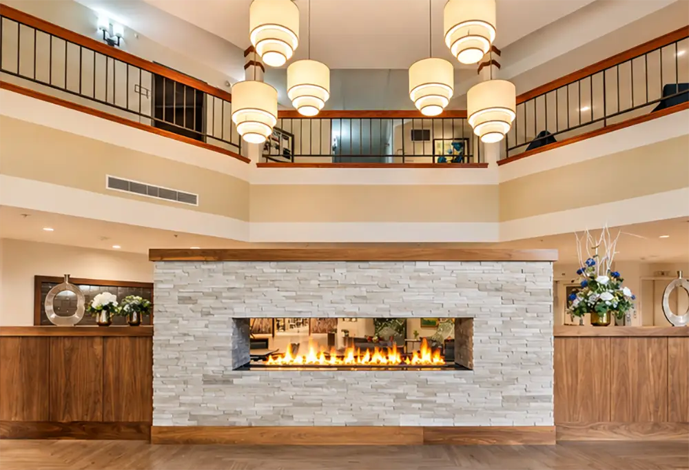 A lobby with a reception desk, modern furnishings, a stone accent wall, chairs, and signage on a stand welcomes guests to Sholom Senior Living.