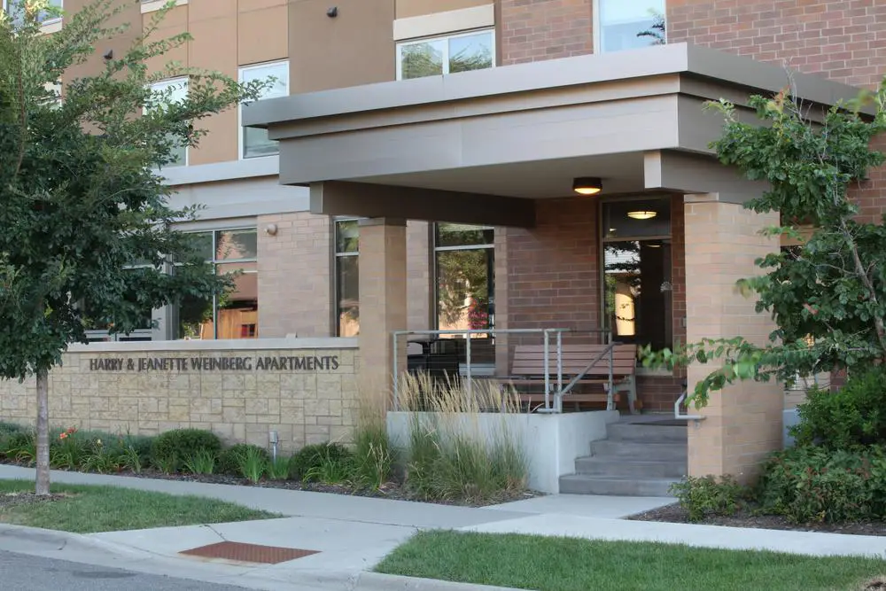 Entrance of a modern Sholom Senior Living apartment building with a sign reading "Harry & Jeanette Weinberg Apartments" beside landscaped greenery and a covered entryway.